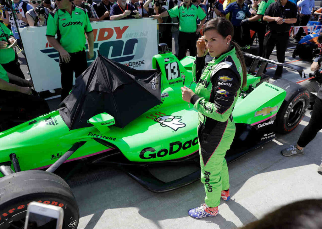Danica Patrick ao lado de seu carro de corrida nos boxes antes de uma prova, vestindo macacão verde e se preparando para a competição. A imagem destaca a trajetória de Danica Patrick como uma das principais referências entre as mulheres no automobilismo.