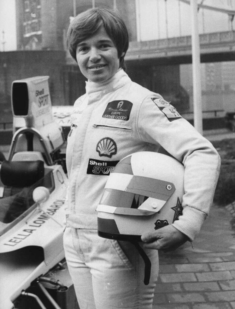 Lella Lombardi, piloto de corrida em pé ao lado de um carro de competição, vestindo macacão e segurando um capacete, durante sessão de fotos nos boxes. A imagem representa a presença histórica das mulheres no automobilismo e sua atuação em categorias de alto nível.