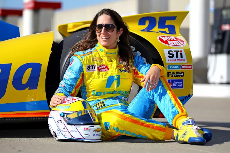 Bia Figueiredo posa ao lado de um carro de corrida, usando macacão amarelo e azul e capacete apoiado no chão. A imagem destaca a trajetória de Bia Figueiredo como uma das principais representantes brasileiras no automobilismo.