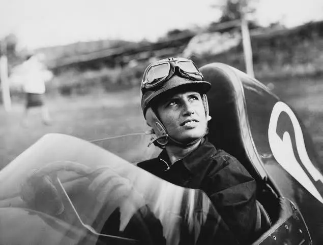 Maria Teresa de Filippis, mulher piloto sentada no cockpit de um carro de corrida antigo, usando capacete e óculos de proteção, olhando para o horizonte antes da largada. A imagem destaca a trajetória das mulheres no automobilismo e sua participação histórica nas competições.