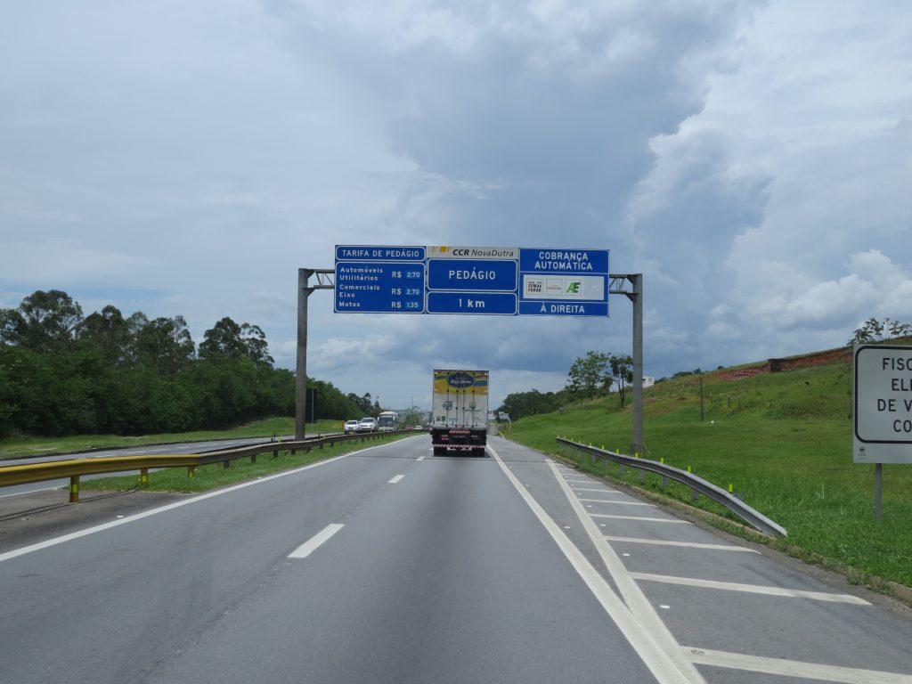 Caminhão trafega pela BR-116 sob placa de sinalização que indica pedágio e cobrança automática na rodovia.