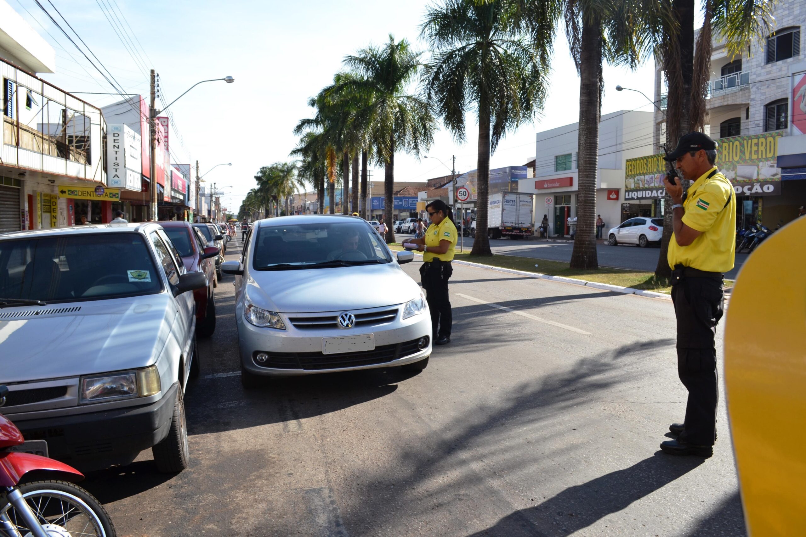 Agentes de trânsito fiscalizam um carro parado em fila dupla ao lado de outros veículos estacionados.